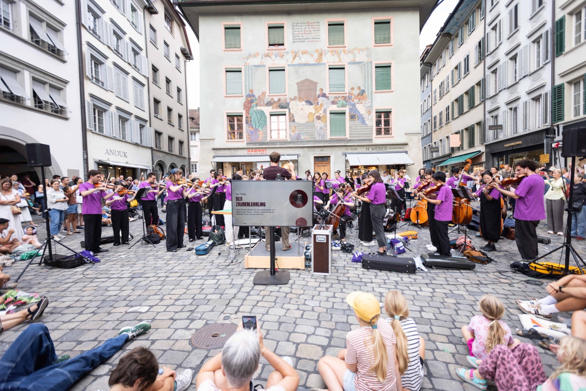 Symphonic Jukebox on Kapellplatz in Lucerne, 2026: young musicians in purple, audience sitting on the square